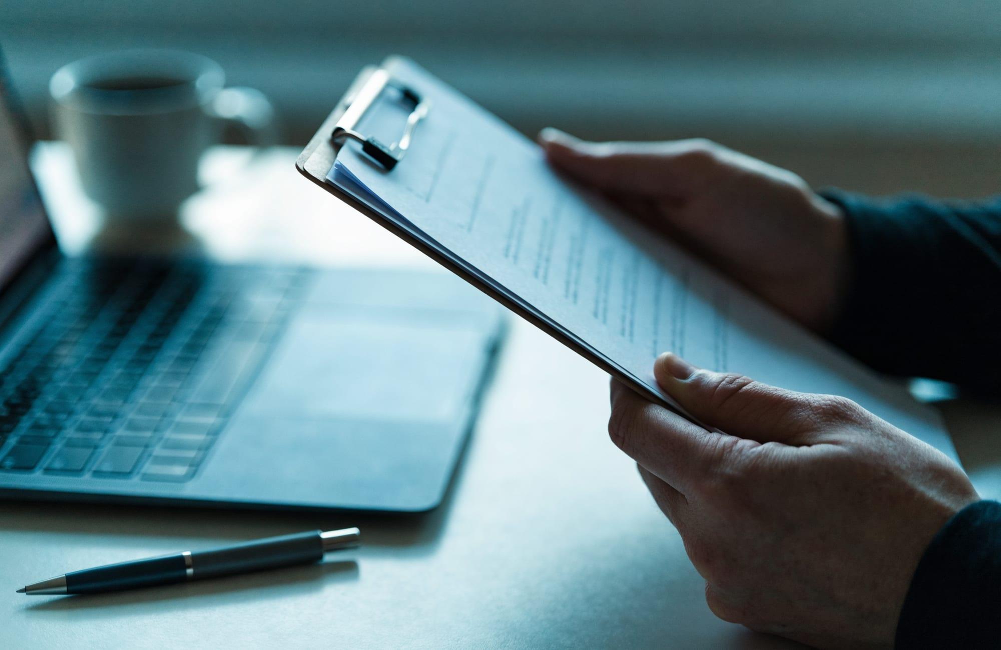Hands holding a clipboard of clinical paperwork beside an open laptop